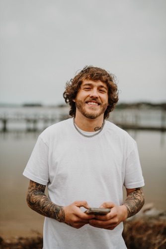 Patrick Moss smiling beside a lake in a white T-shirt, holding a phone with both hands.