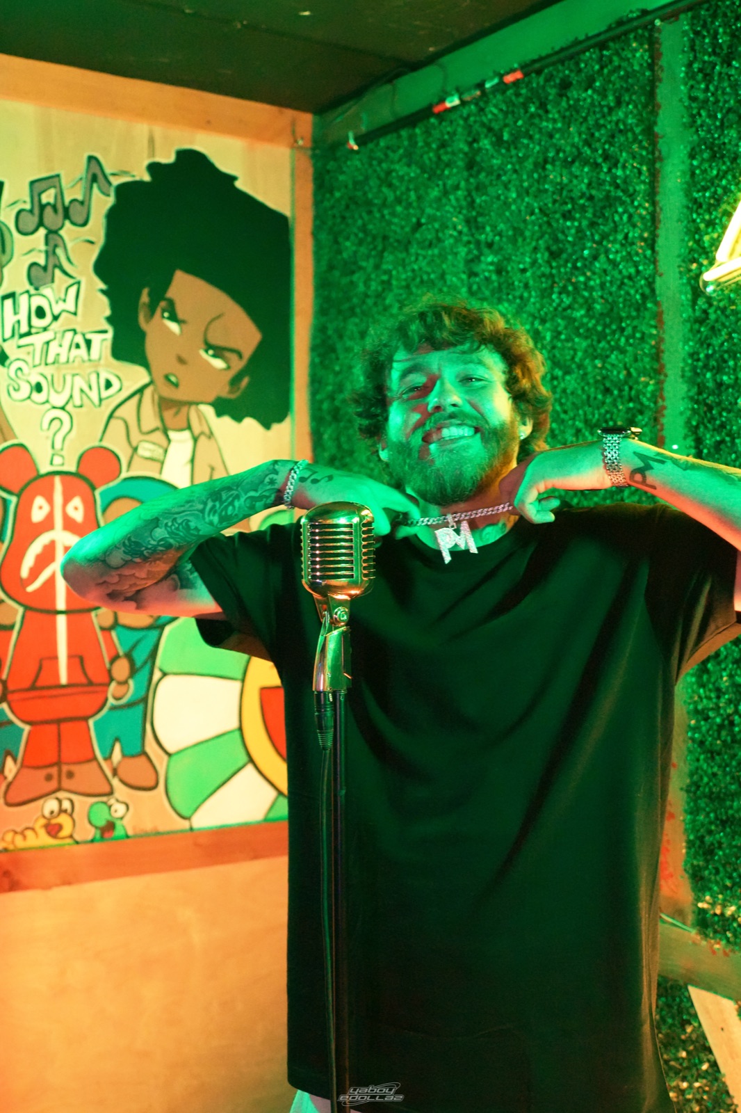 Patrick Moss holding his PM pendant beside a gold microphone under green lighting.