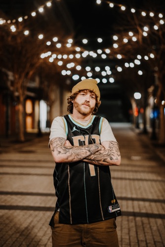 Patrick Moss standing with his arms crossed beneath string lights, wearing a tan beanie and black basketball jersey.