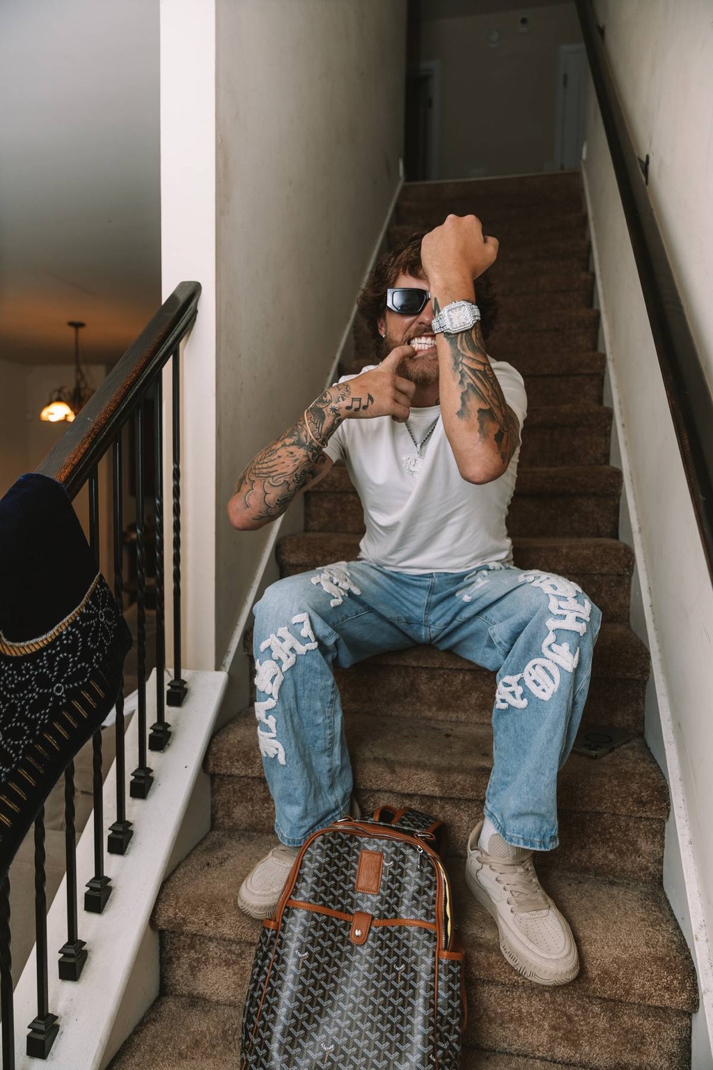 Patrick Moss seated on a staircase in a white T-shirt and jeans, raising his diamond watch and showing his grin.