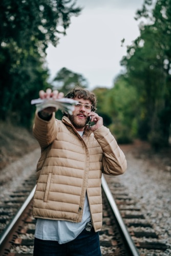 Patrick Moss on railway tracks in a tan puffer jacket, holding a phone to his ear and cash toward the camera.